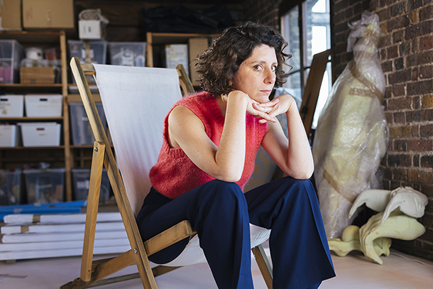 A woman sitting on a folding canvas and wooden chair in an artist studio. The room has a brick wall as well as many art materials and supplies in the background.